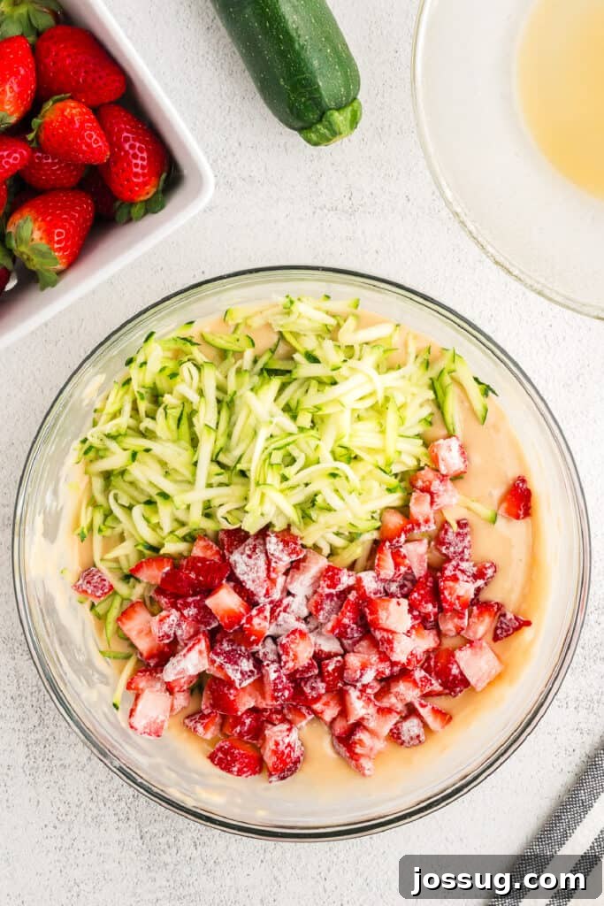 The bowl of strawberry zucchini bread batter is now topped with a generous layer of vibrant grated zucchini and diced, floured strawberries, ready to be gently folded in.