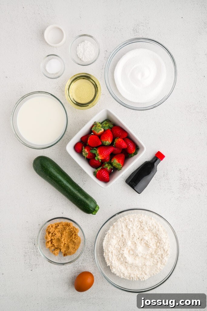 An overhead shot showcasing all the fresh ingredients for Strawberry Zucchini Bread meticulously arranged on a clean white surface, highlighting colorful strawberries and grated zucchini.