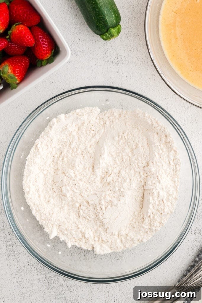 The dry ingredients are thoroughly mixed in their bowl, with fresh, vibrant red strawberries and green grated zucchini displayed alongside, awaiting their turn.