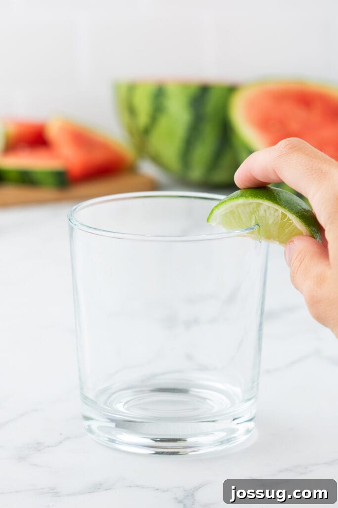 Crimson Wave Margarita 4 Hand rubbing a lime wedge along the rim of a clear glass with watermelon in the background to prepare for salting