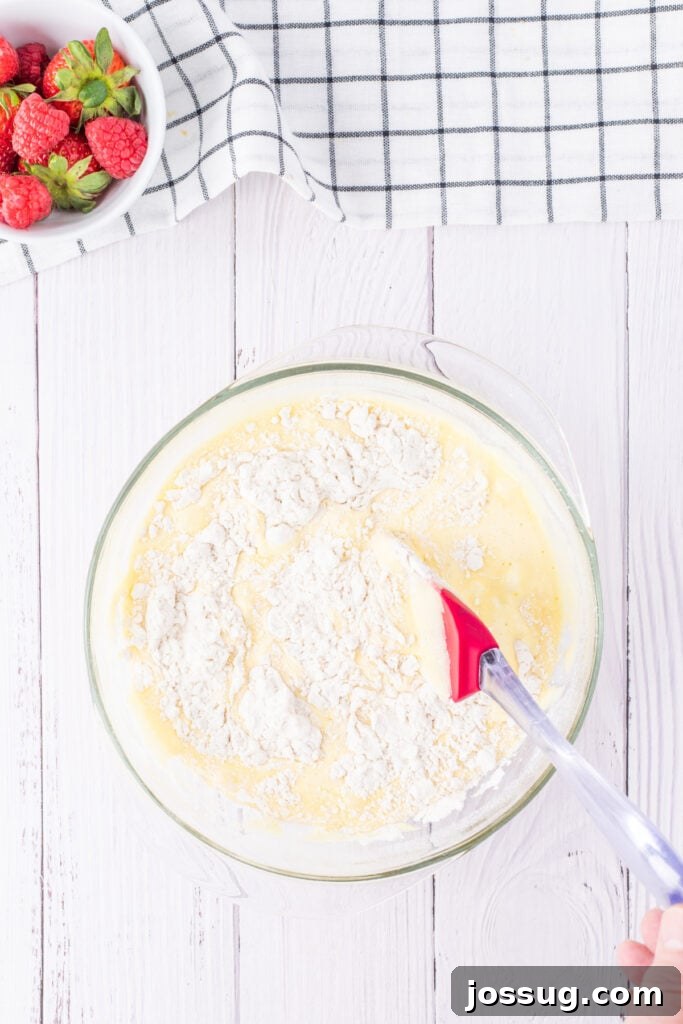 Creamy Three Milk Dream 5 Dry cake ingredients, including flour, baking powder, and salt, being gently folded into the airy egg yolk mixture within a large glass bowl using a red spatula, illustrating a key step in preparing the Tres Leches cake batter.