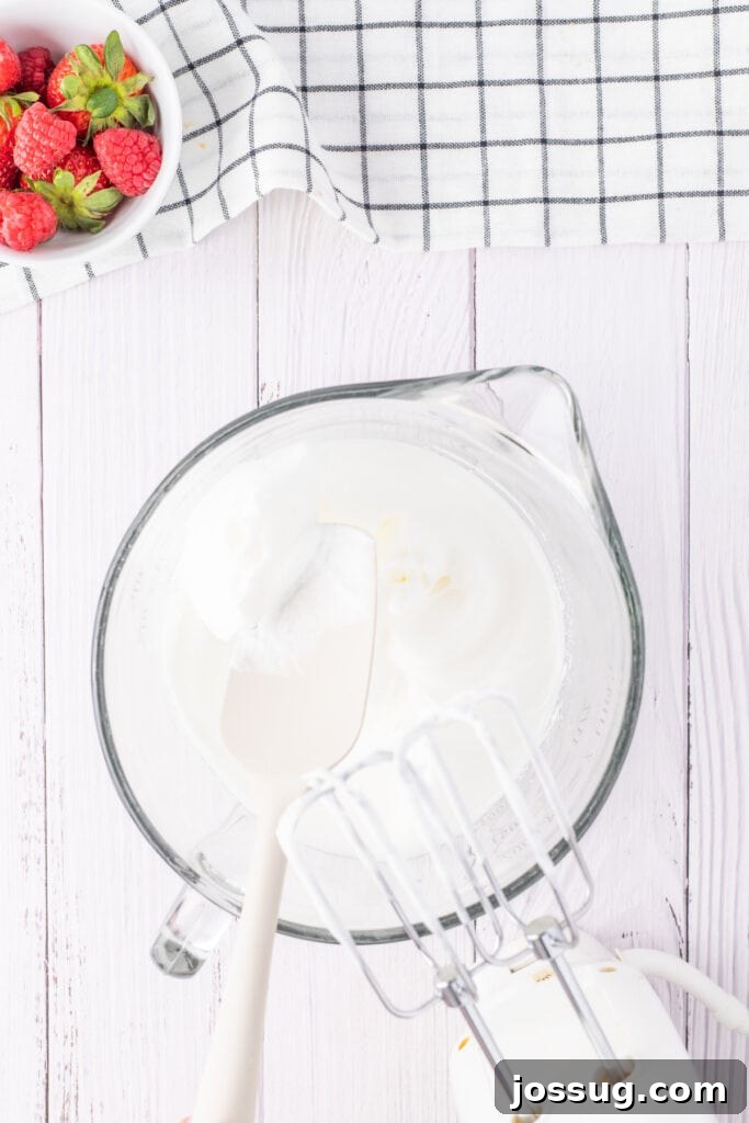 Creamy Three Milk Dream 6 Heavy whipping cream being vigorously mixed in a clear glass bowl using an electric hand mixer, creating fluffy peaks for the delicious whipped topping of the Tres Leches cake.