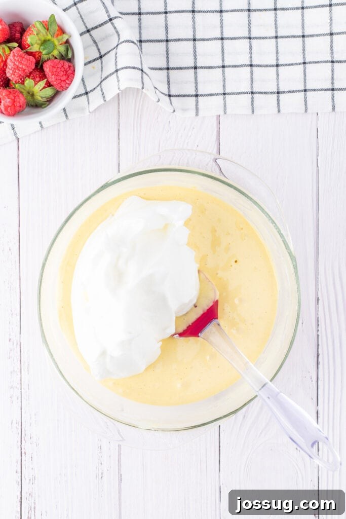 Creamy Three Milk Dream 7 Delicate whipped egg whites are being carefully folded into the main cake batter in a large glass bowl with a red spatula, demonstrating the gentle technique required to maintain airiness for the perfect Tres Leches sponge.