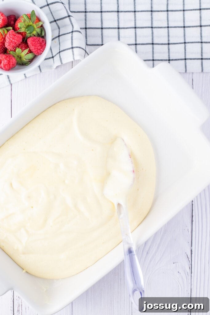 Creamy Three Milk Dream 8 Raw cake batter is spread evenly across the bottom of a pristine white ceramic baking dish using a spatula, smoothed out and ready to be placed into the oven for baking.