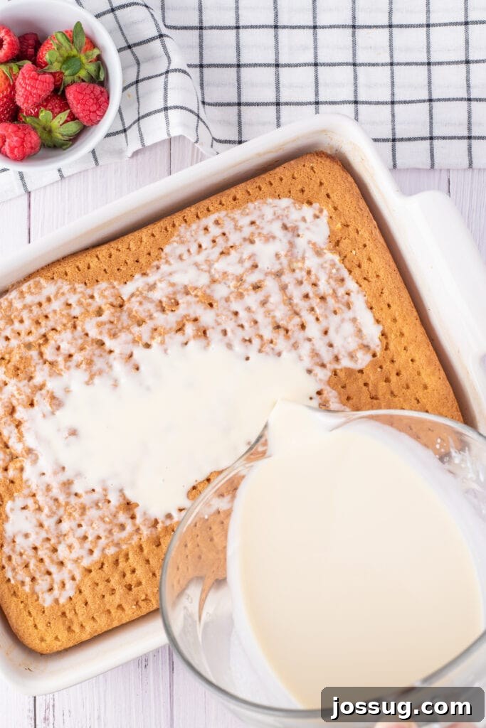 Creamy Three Milk Dream 9 The luxurious Tres Leches milk mixture is being slowly and evenly poured over the surface of the freshly baked and poked cake in a white baking dish, ensuring thorough absorption. A bowl of glistening fresh mixed berries sits nearby, ready for garnish.