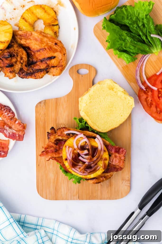 A beautifully layered Grilled Hawaiian chicken sandwich with bacon, pineapple, red onion, and a drizzle of BBQ sauce, served on a cutting board.