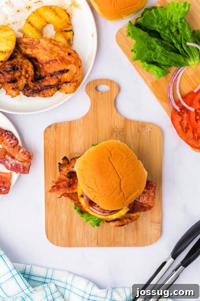 A fully assembled grilled Hawaiian chicken sandwich with fresh lettuce, tomato, juicy chicken, crispy bacon, sweet pineapple, and sharp red onion on a soft bun.