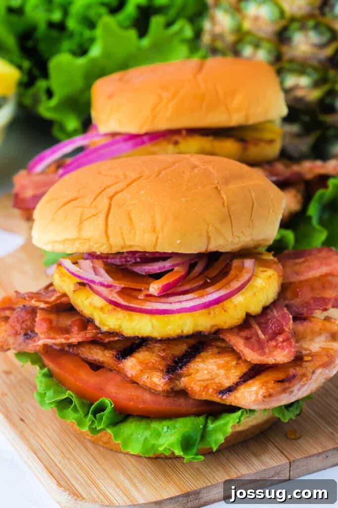 Close-up of a stacked grilled Hawaiian chicken sandwich showing layers of chicken, bacon, pineapple, and red onion, highlighting its delicious construction.