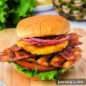 Square format image of a grilled Hawaiian chicken sandwich piled high with lettuce, tomato, bacon, pineapple, and red onion on a soft bun.