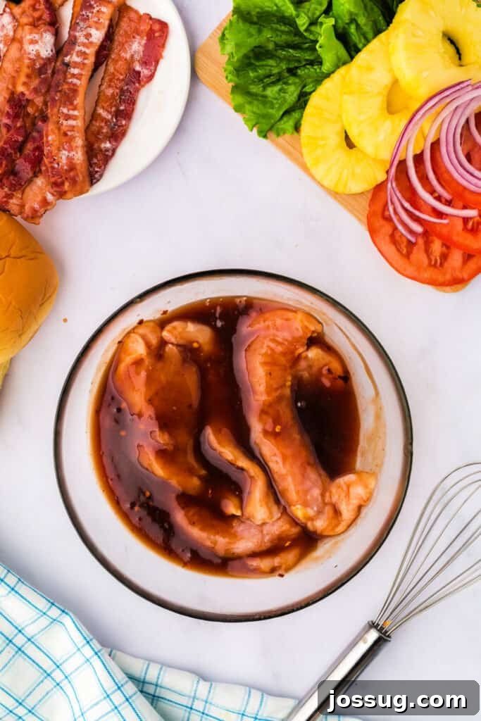 Chicken breasts marinating in the pineapple BBQ sauce mixture, absorbing flavors, with surrounding sandwich ingredients prepped.