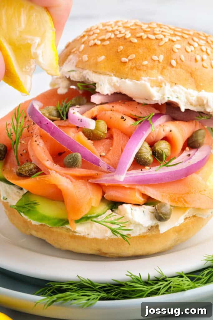 Close-up of a sesame bagel sandwich being finished with a squeeze of lemon over smoked salmon, red onions, and capers.