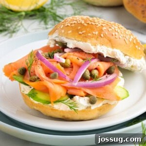 Square image of a smoked salmon bagel sandwich with cream cheese, red onions, capers, dill, and cucumber on a sesame bagel.