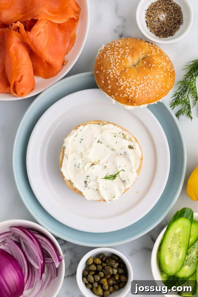 Sesame bagel half spread with herbed cream cheese surrounded by ingredients like smoked salmon, cucumber, red onion, and capers.