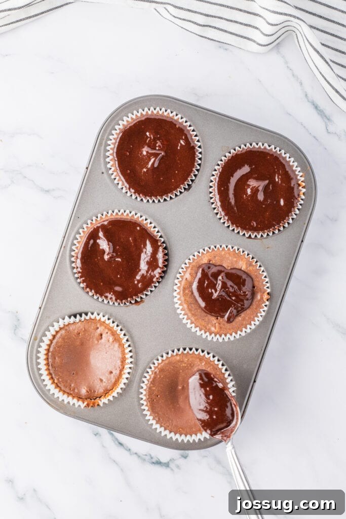 Spoon spreading ganache onto baked mini chocolate cheesecakes in a muffin pan
