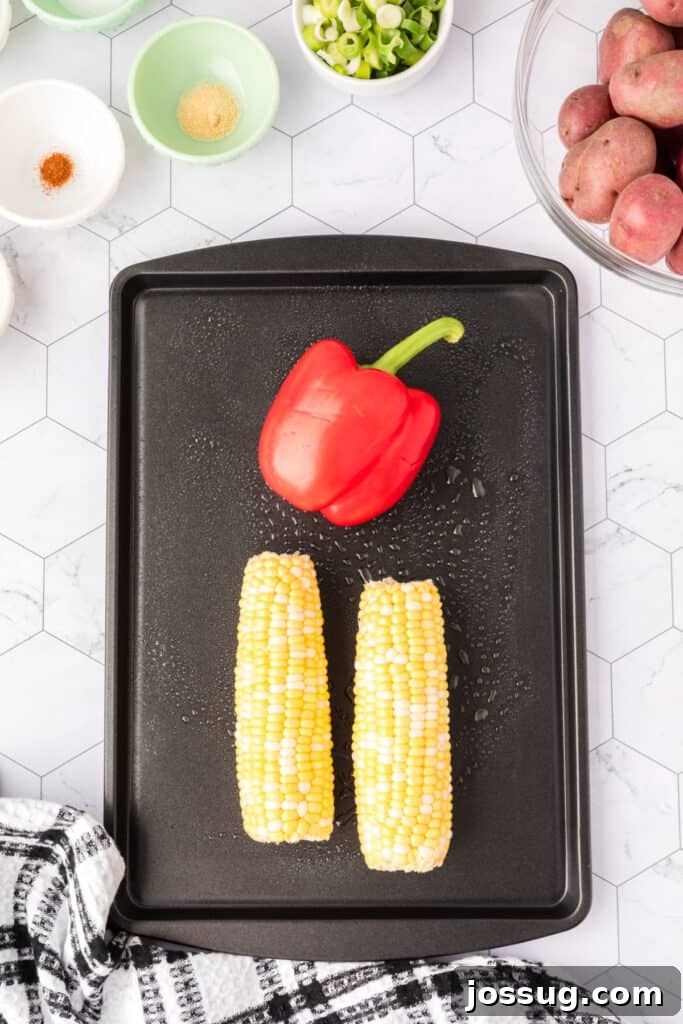 Red bell pepper and two ears of corn placed on a black baking sheet, ready to roast.
