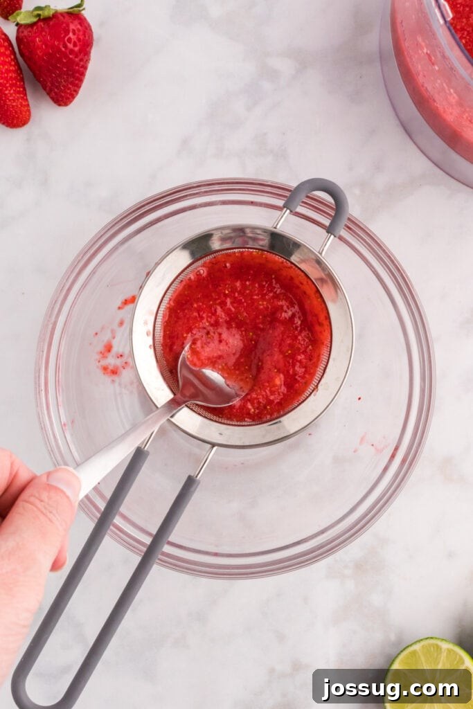 Blush Burst Cooler 6 Strawberry purée being strained through a fine mesh strainer with a spoon over a glass bowl to remove seeds and pulp.