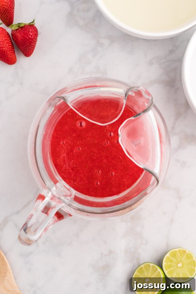 Blush Burst Cooler 9 Strawberry purée being poured into the lime juice and sugar mixture in a glass pitcher, beginning the blending process.