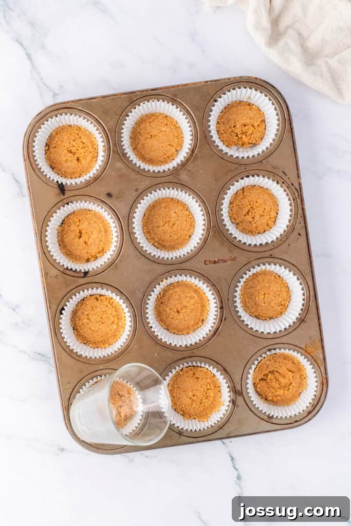 Effortless Chocolate Peanut Butter Cheesecake Bites 3 Muffin pan lined with white cupcake liners, each filled with pressed graham cracker crust. A small glass is shown pressing the crust into shape.