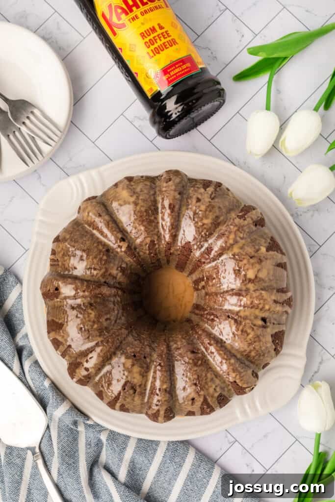 Decadent Kahlua Chocolate Bundt 13 The magnificent finished Chocolate KahlĂșa Bundt Cake, generously coated with a glistening glaze, elegantly presented alongside a KahlĂșa bottle and a vase of delicate white tulips, creating a truly inviting scene.