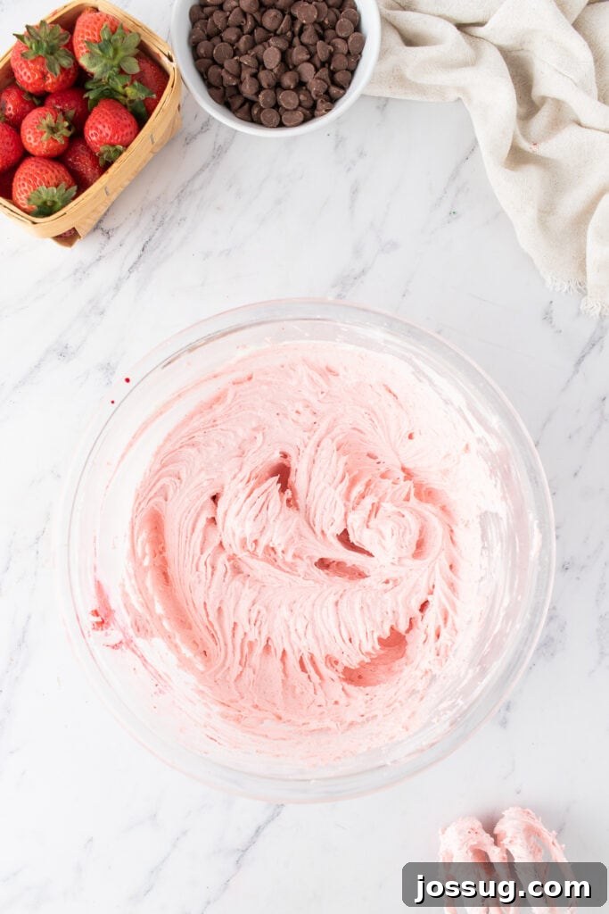 Bowl of fluffy pink strawberry buttercream frosting with a hand mixer and fresh strawberries nearby.
