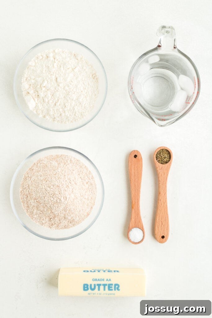 Overhead view of ingredients for galette crust including whole wheat and all-purpose flours, cold butter, spices, and iced water, laid out on a clean surface.