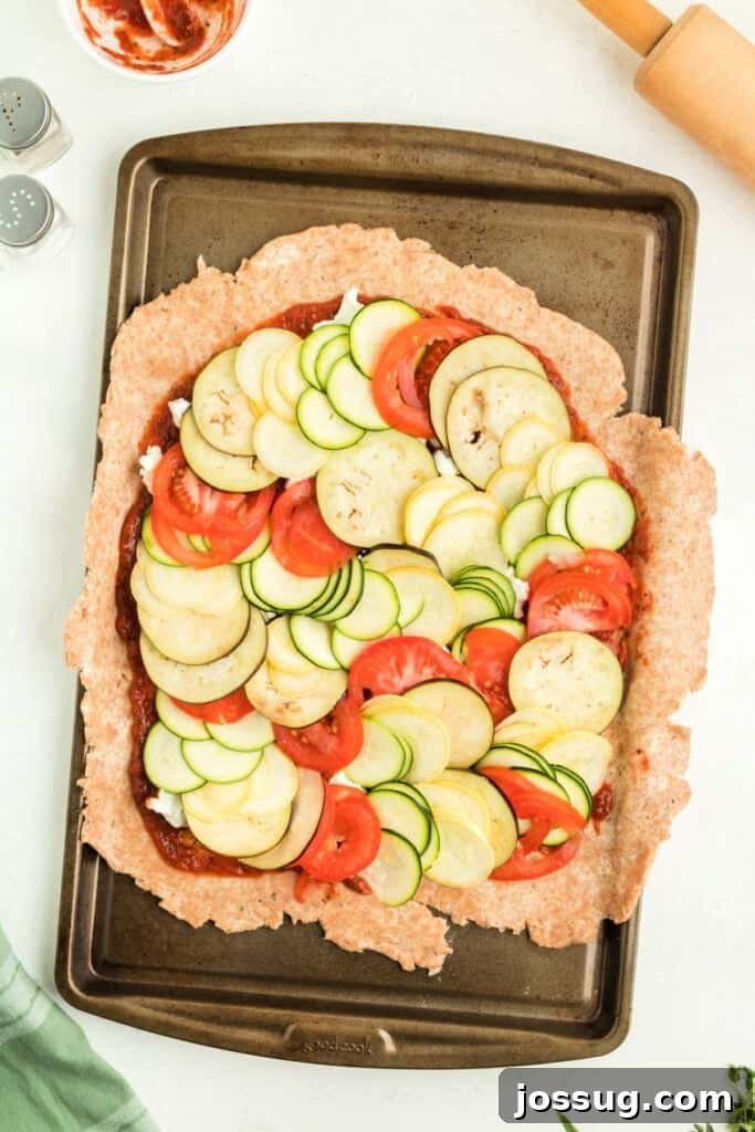 Unbaked galette crust spread on parchment paper on a baking sheet, generously topped with marinara sauce, crumbled goat cheese, and artfully layered vegetables.