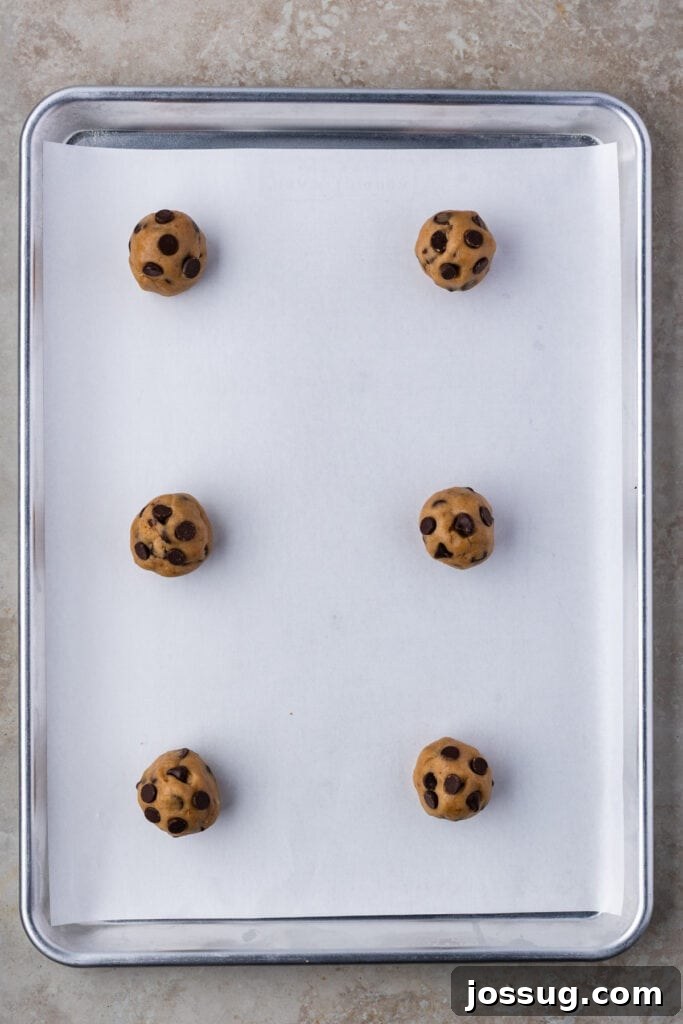 Six perfectly scooped and uniformly sized balls of chocolate chip cookie dough, neatly spaced on a parchment-lined baking sheet, awaiting their turn in the oven.