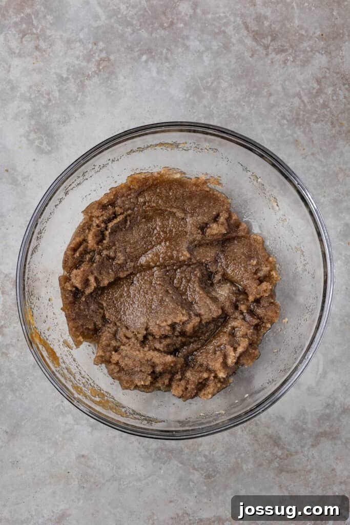 A mixture of rich brown sugar and granulated sugar smoothly combined with the cooled brown butter in a clear glass mixing bowl, exhibiting a cohesive, creamy texture.