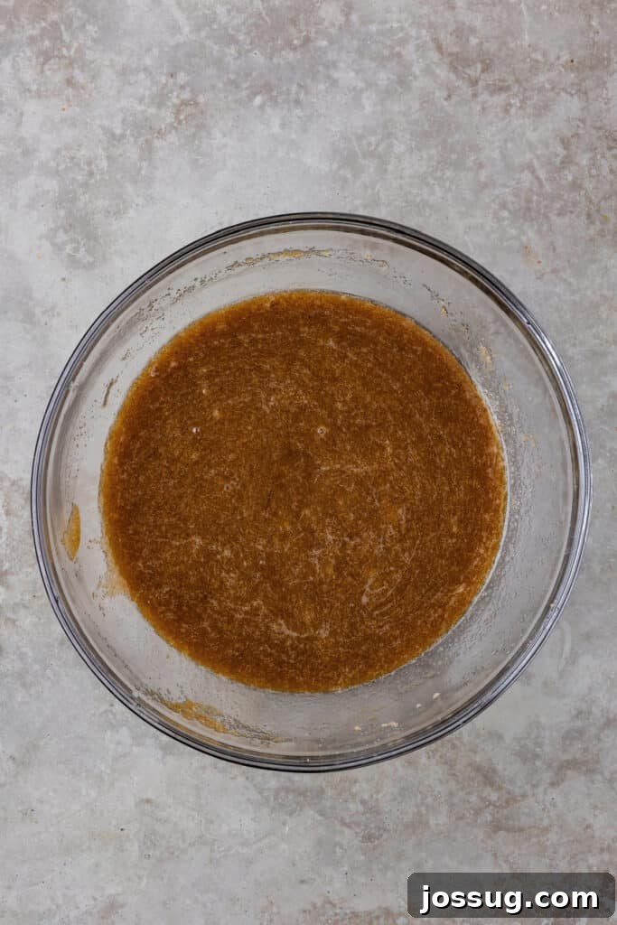 The wet ingredients, including brown butter, sugars, eggs, and vanilla, perfectly whisked together in a glass bowl, creating a smooth, light, and glossy batter.
