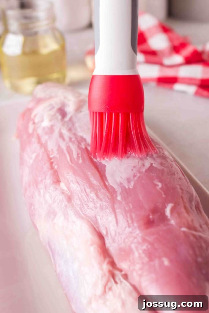 Olive oil being brushed onto raw pork tenderloin using a red silicone brush