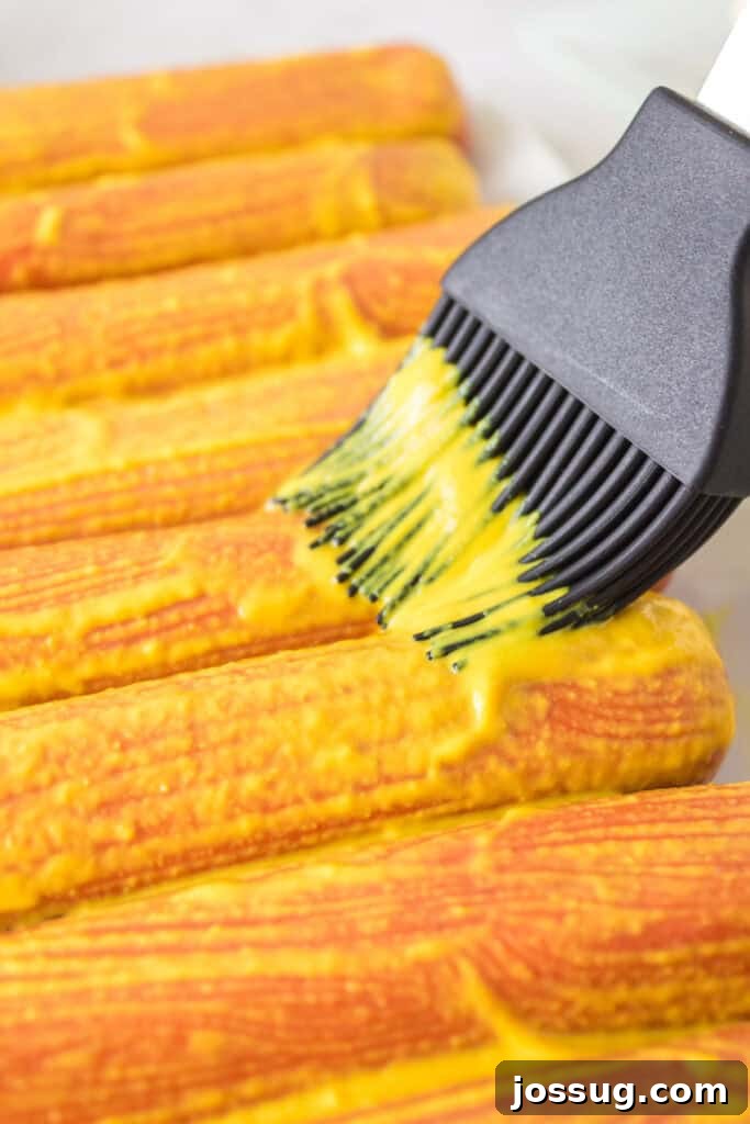 Close-up of a basting brush spreading yellow mustard onto hot dogs before seasoning