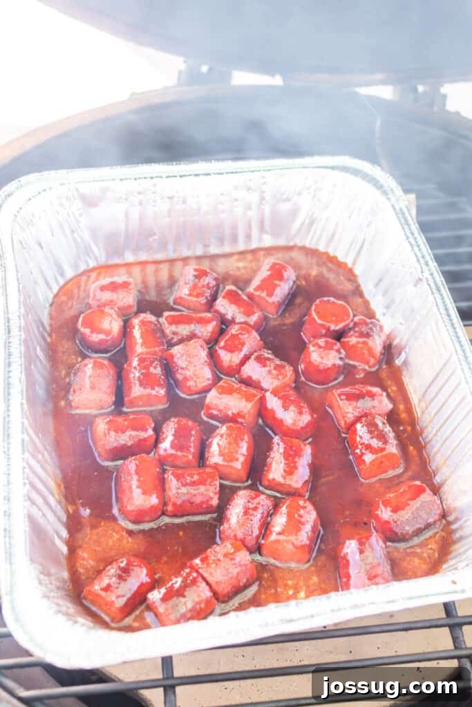 Aluminum tray of saucy hot dog bites sitting on a smoker grill with smoke rising