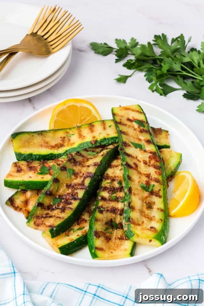 Side angle of grilled parmesan zucchini served on a white plate with fresh parsley and lemon. The zucchini slices have visible golden-brown grill marks and melted parmesan cheese, garnished for freshness.