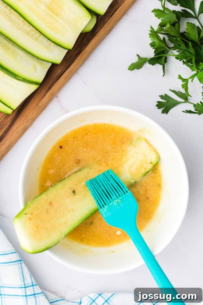 A zucchini slice being generously brushed with the parmesan breadcrumb butter mixture using a blue basting brush.