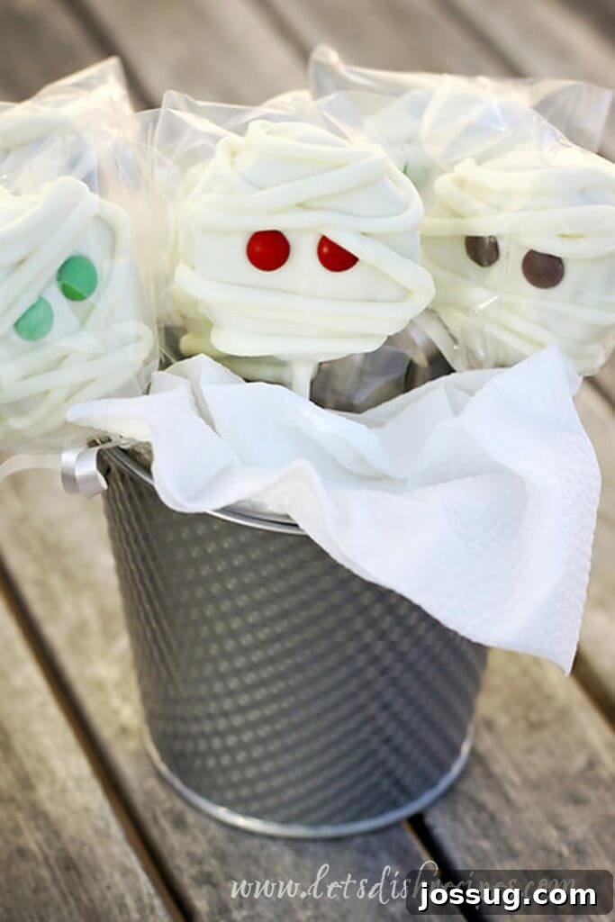Cute mummy pops made from cake or cookie dough, decorated with white icing and edible eyes, displayed in a tin pail.