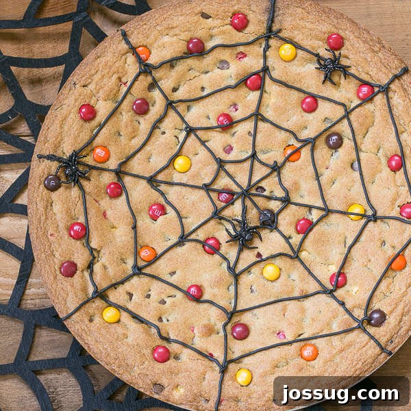 A large, round chocolate chip cookie cake decorated with white frosting in a spiderweb pattern, resting on a cutting board.
