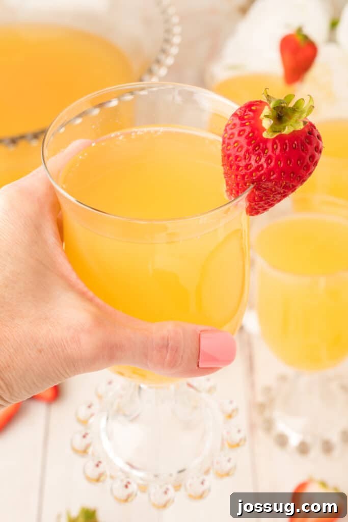 Sparkling Celebration Punch 5 Hand holding a glass of mock champagne punch with a fresh strawberry on the rim and punch bowl in the background.
