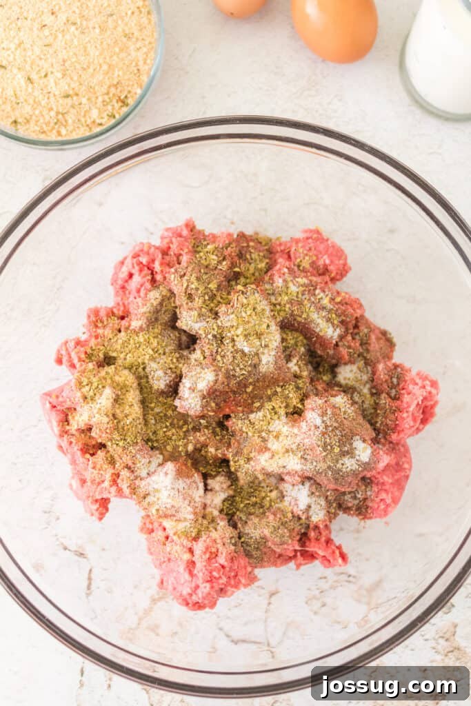 Pit Smoked Meatloaf 4 A vibrant mix of seasonings being generously sprinkled over ground beef in a clear glass mixing bowl, signaling the start of flavor infusion.