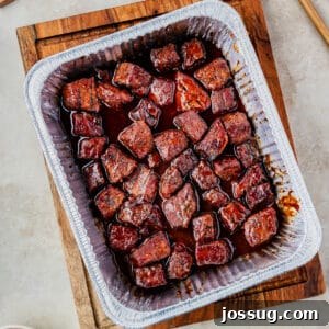 Overhead view of BBQ-glazed Poor Man’s Burnt Ends in a foil pan on a wooden board, square crop, ready to be enjoyed.