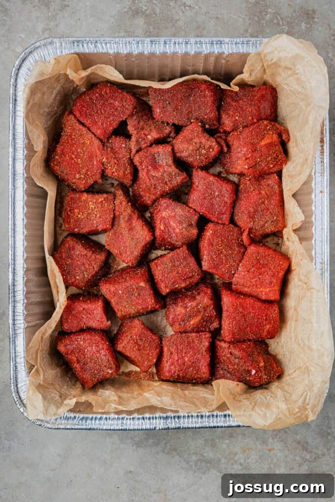 Burnt Ends on a Budget 5 Chuck roast cubes coated evenly in dry rub, arranged in a foil pan lined with parchment paper, prepared for smoking.