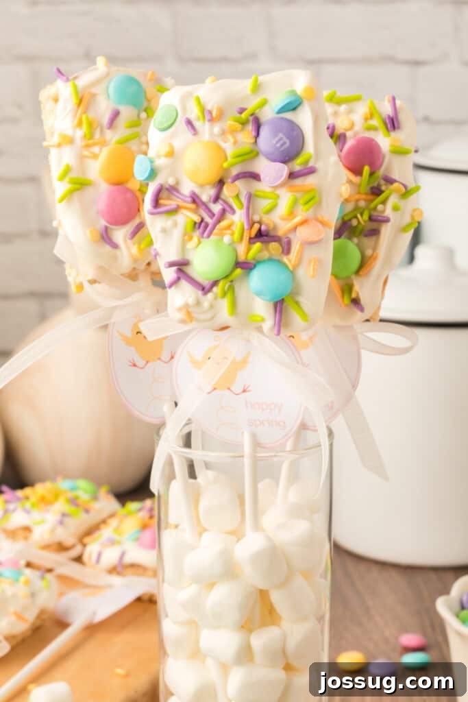 Three colorful Spring Rice Krispies Treats Pops displayed in a glass cylinder filled with mini marshmallows, ready for a festive spring celebration.