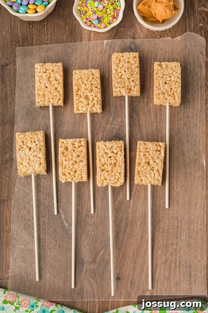 Unwrapped Rice Krispies treats carefully skewered onto long lollipop sticks, resting neatly on a sheet of waxed paper, prepared for the next coating steps.