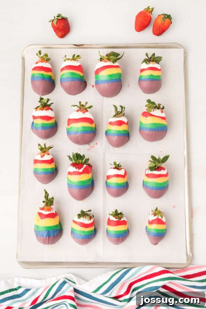 A baking sheet lined with parchment paper, holding several freshly candy coated strawberries in rainbow colors.