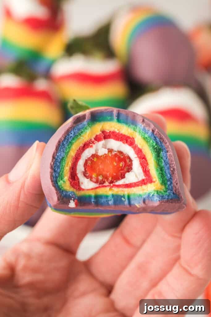 A hand holding a perfectly coated rainbow strawberry, cut in half to show the distinct, colorful layers of candy coating over the fresh strawberry.