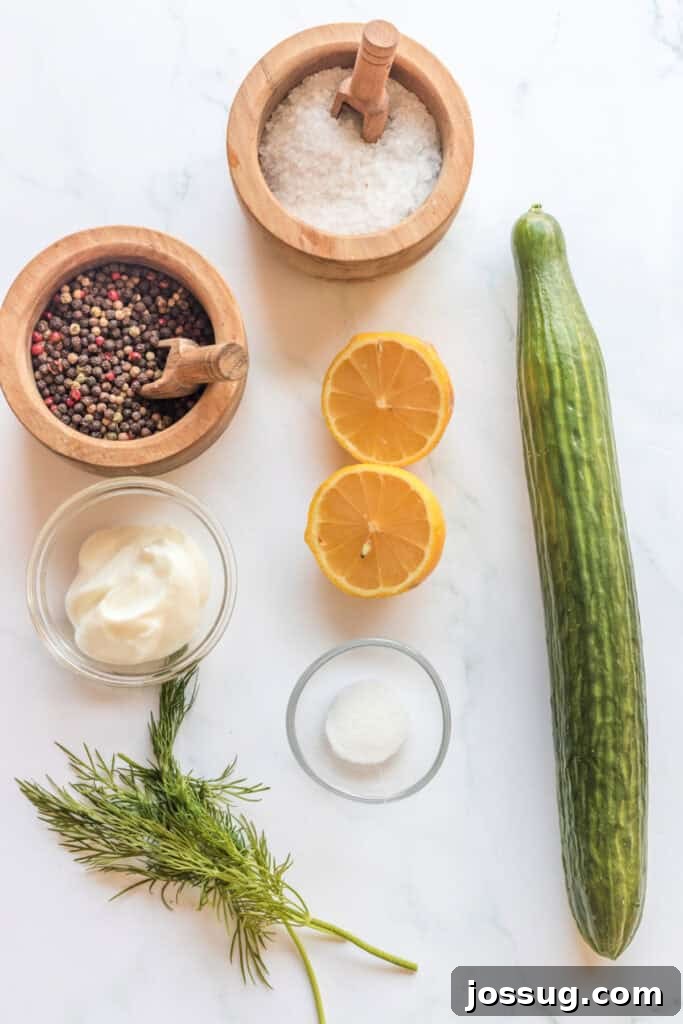 Polish Dill Cucumber Salad 3 Key ingredients for Polish cucumber salad laid out on a white marble countertop, including a fresh lemon, a bunch of dill, a tub of sour cream, a whole English cucumber, salt, granulated sugar, and black peppercorns.