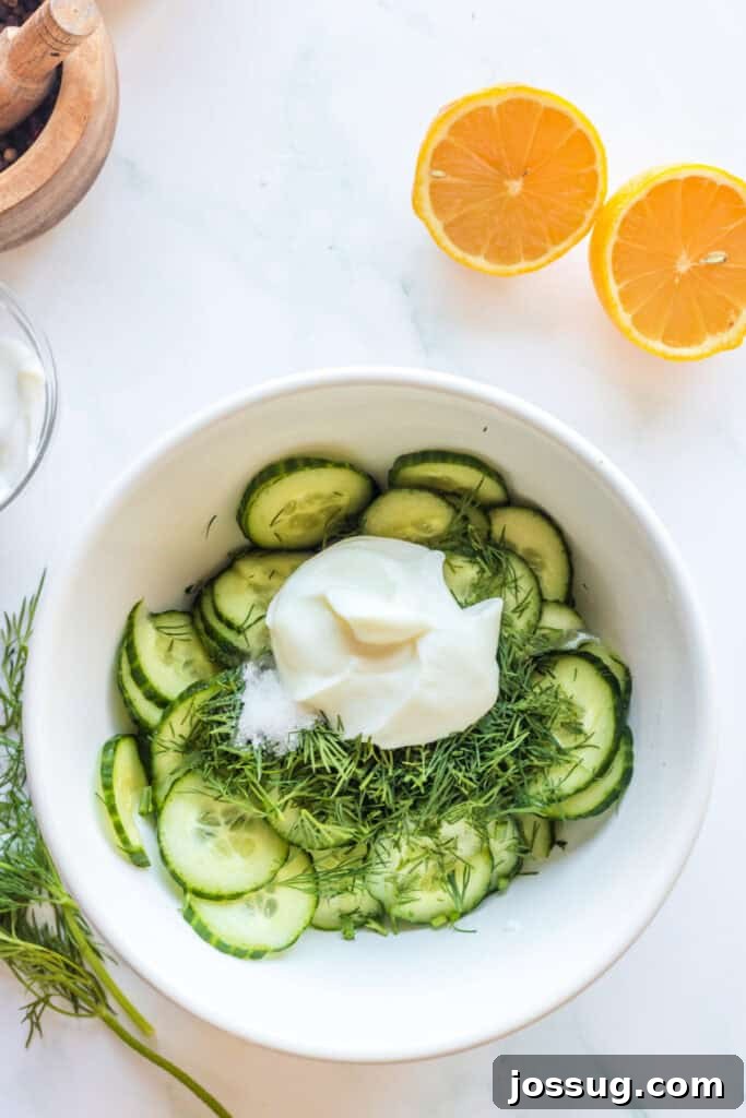 Polish Dill Cucumber Salad 6 A large white bowl containing drained cucumber slices, topped with fresh dill, creamy sour cream, salt, and lemon components, poised for mixing into the dressing.