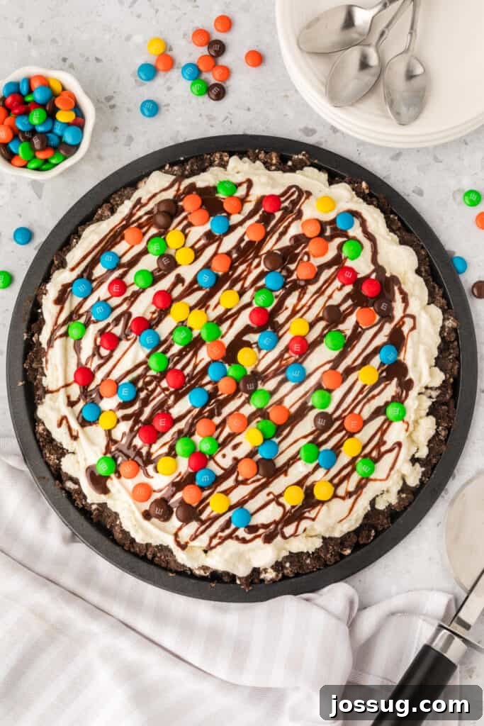 Dessert Pizza Dreams 2 Overhead view of a vibrant ice cream pizza generously topped with colorful M&M's and rich chocolate drizzle, resting on a sleek black pizza pan, ready to be sliced and served.