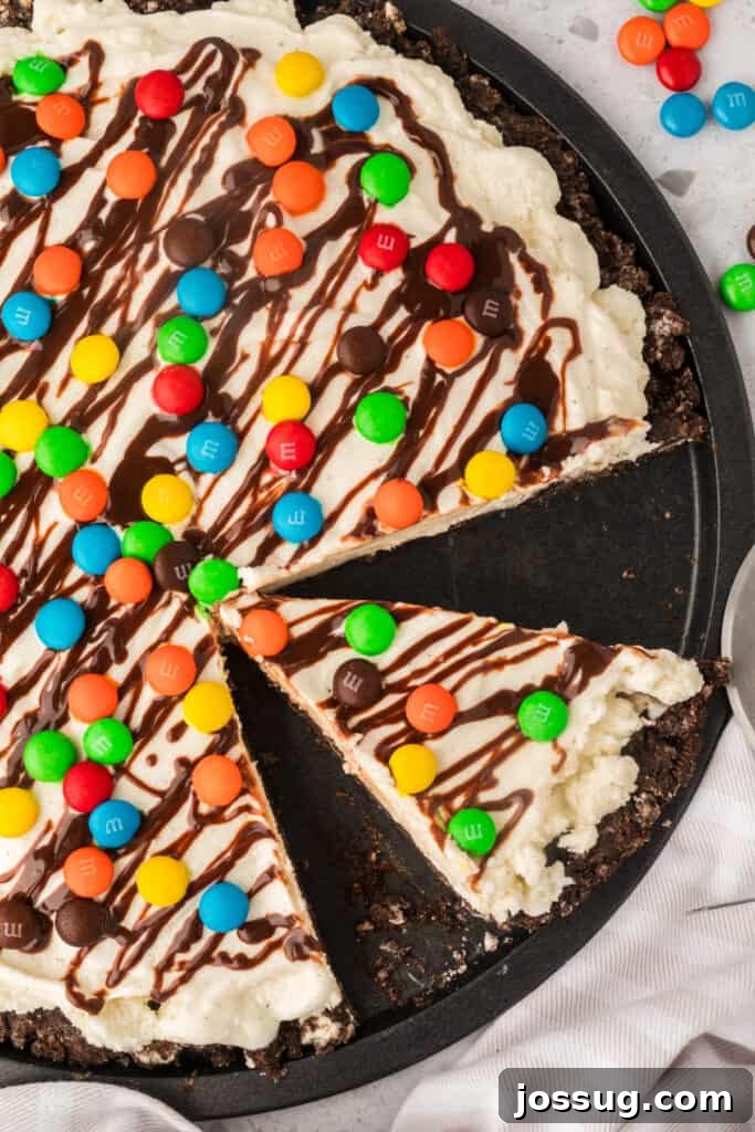Dessert Pizza Dreams 4 An inviting overhead shot of a partially sliced ice cream pizza, with two wedges removed, revealing its delicious layers of cookie crust, vanilla ice cream, hot fudge, and M&M's, all resting on a black pizza pan.