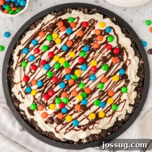A perfectly square crop of a vibrant ice cream pizza featuring a crisp chocolate cookie crust, a thick layer of creamy vanilla ice cream, generous chocolate fudge drizzle, and a colorful scattering of M&M candies, all ready to be enjoyed.
