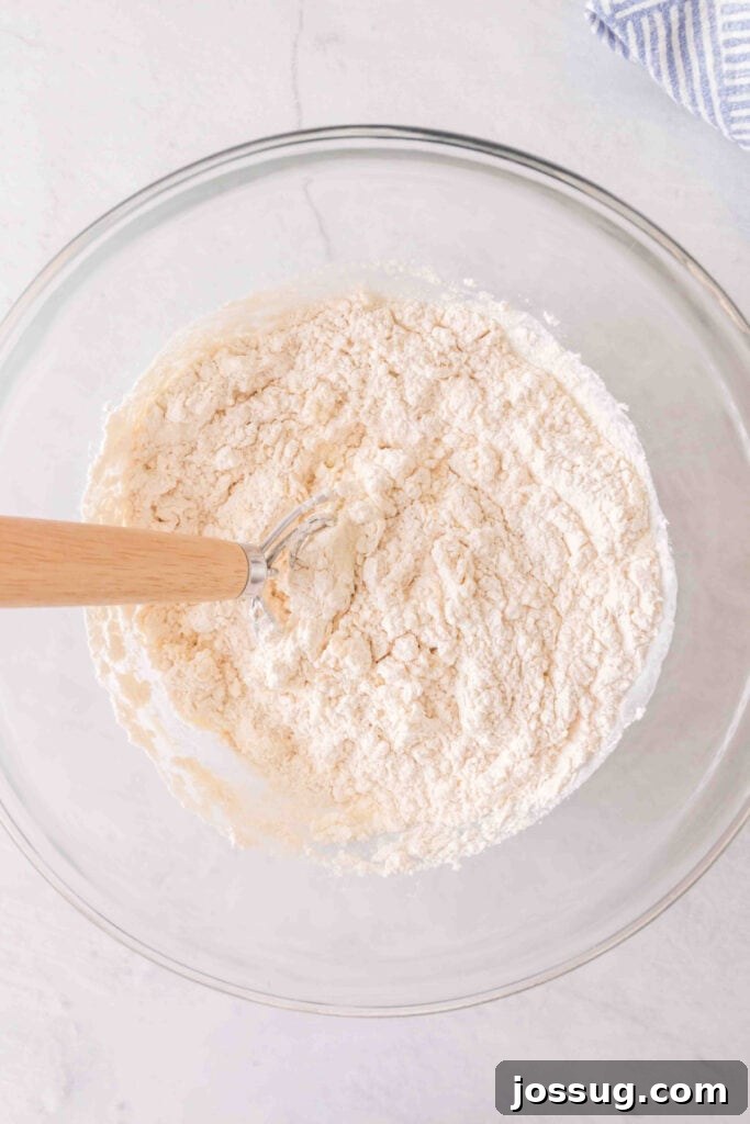 Amazing 3 Ingredient Scones 5 Close-up of scone dough partially mixed in a bowl with a rustic Danish dough whisk, showing the thick, shaggy texture of the ingredients coming together.